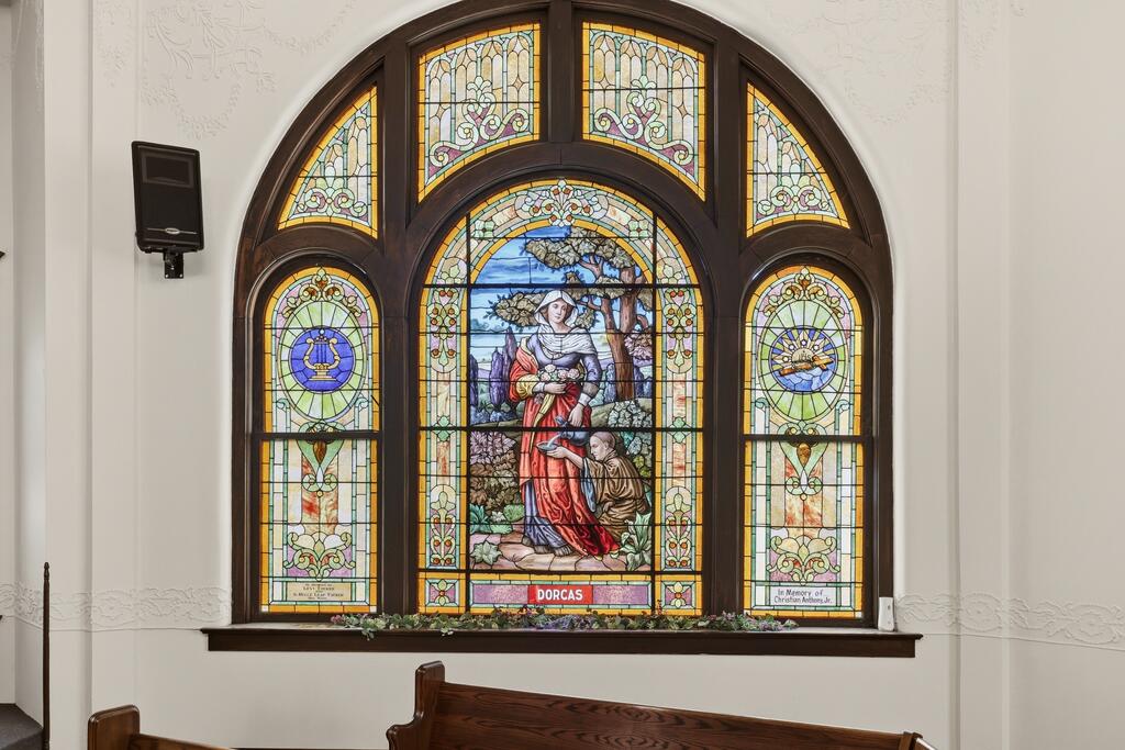 Arched stained-glass window depicting two figures, with a wooden bench below.