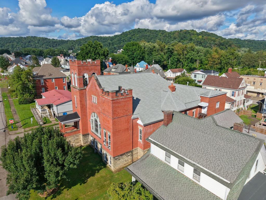Aerial view of a red brick building with a gray roof among town homes and wooded hills.