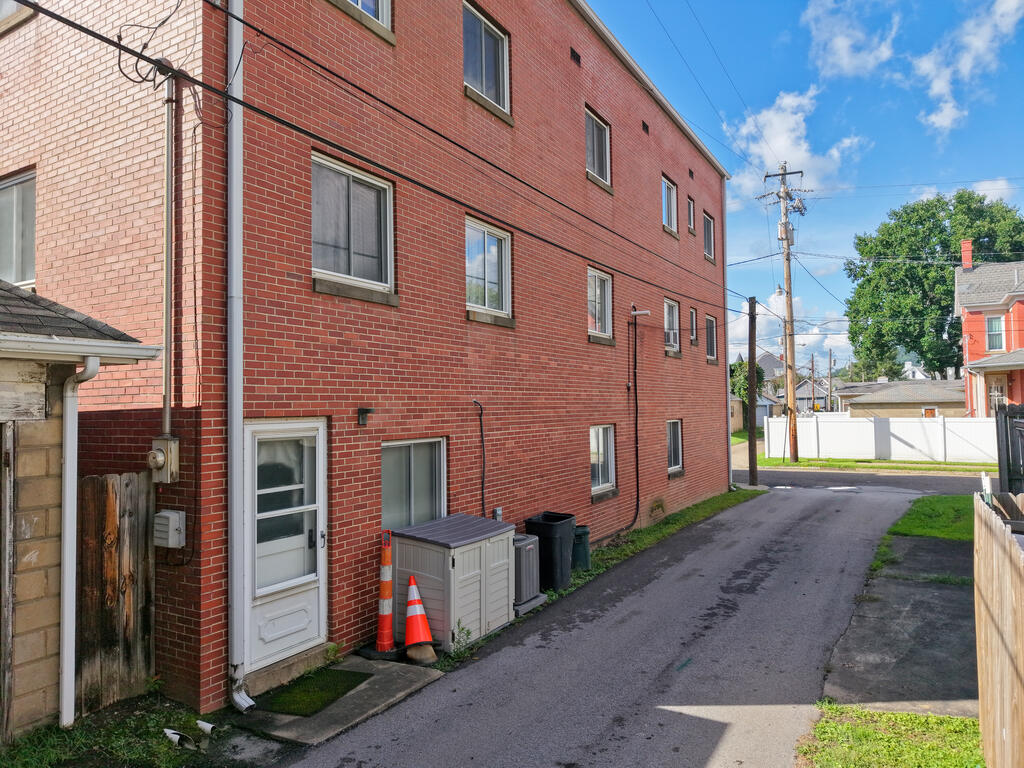 Red-brick multi-story building beside a paved alley, with a white ground-level door and utility bins.