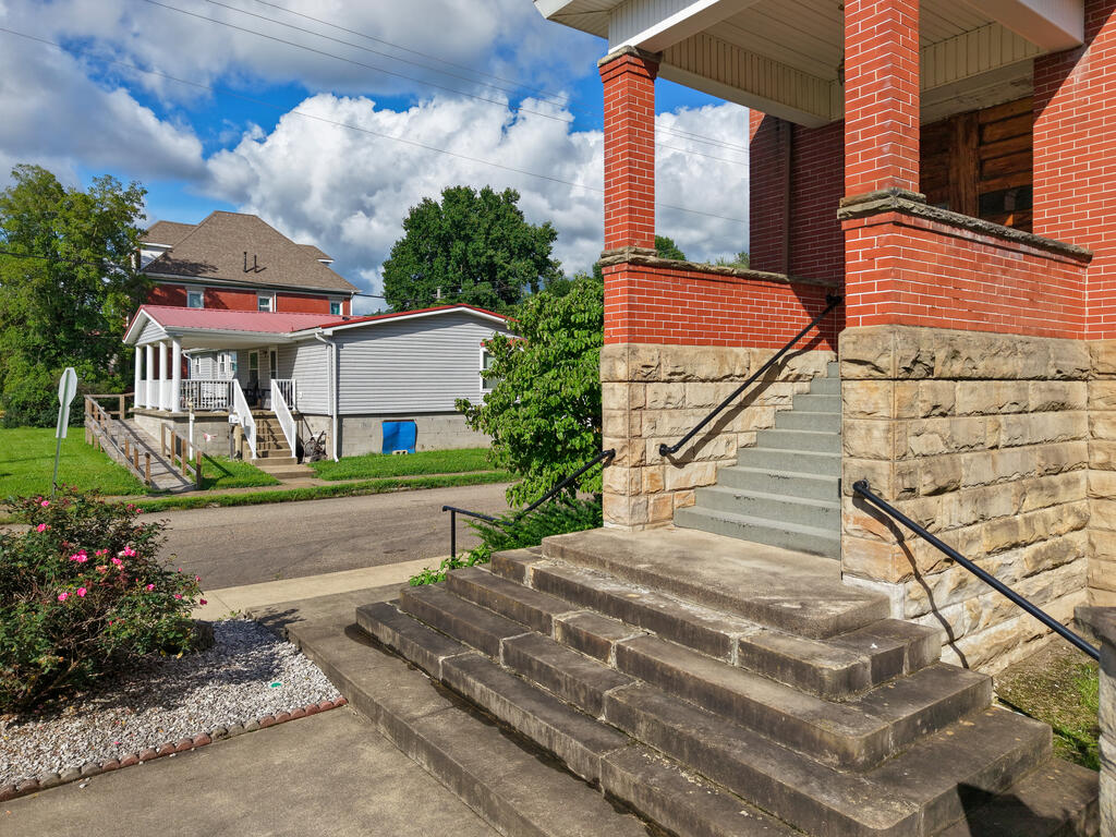 Brick-and-stone building entrance with broad steps and black handrails, across from a white-sided house.