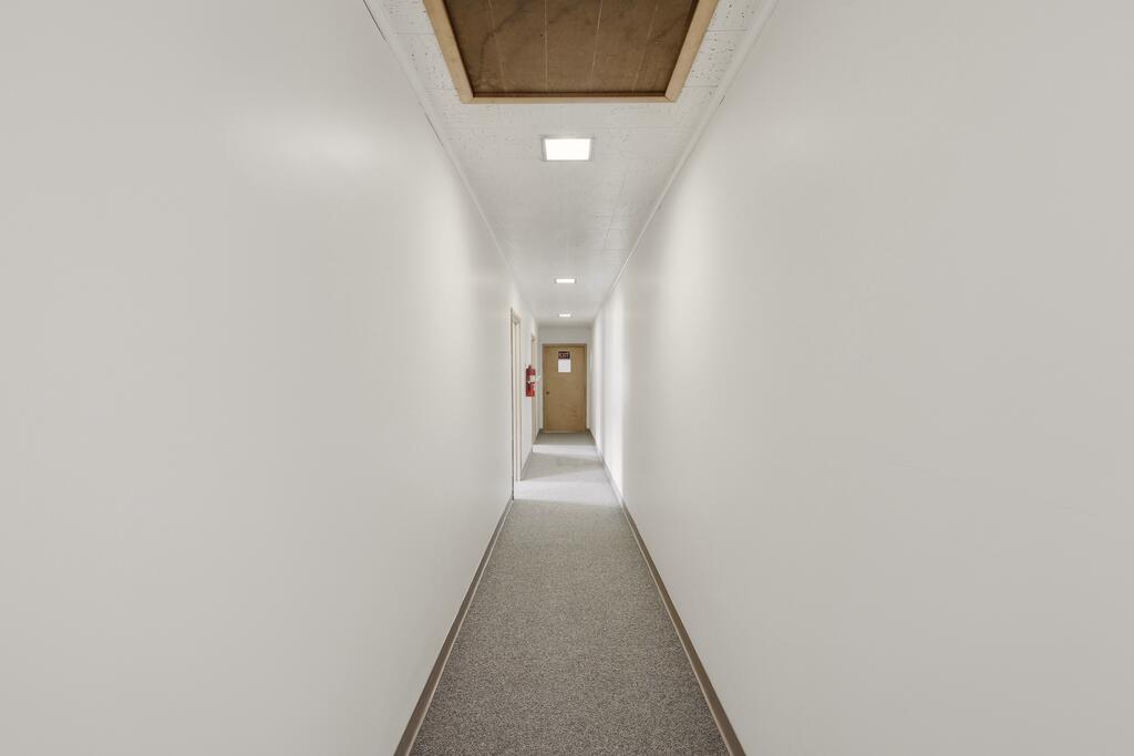 Narrow white hallway with gray carpet, fluorescent lights, and a wooden door at the far end.