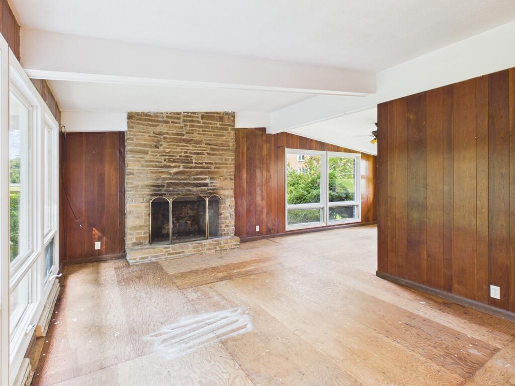 Vacant living room with a stone fireplace, wood-paneled walls, and large windows.