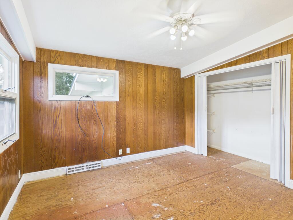 Empty bedroom with wood-paneled walls, a ceiling fan, an open closet, and a worn wood floor.