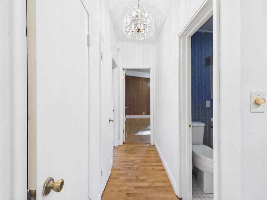 White hallway with wood flooring and a chandelier, with an open doorway to a blue-wallpapered bathroom.