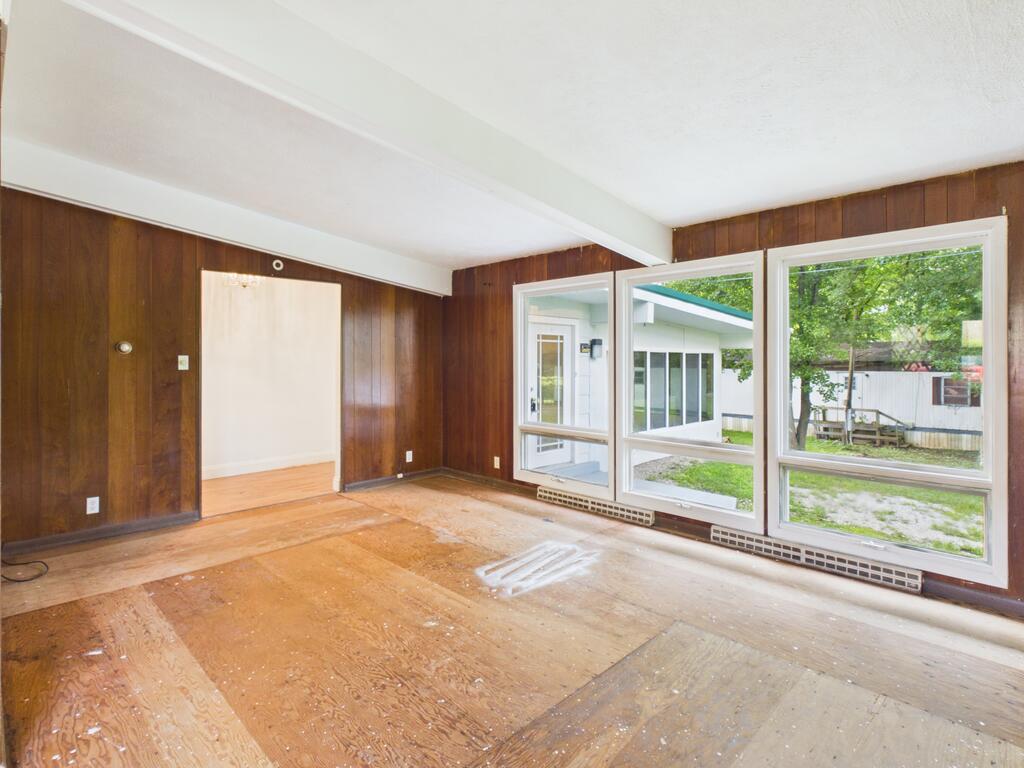 Empty wood-paneled room with a bare plywood floor and large windows overlooking the yard.