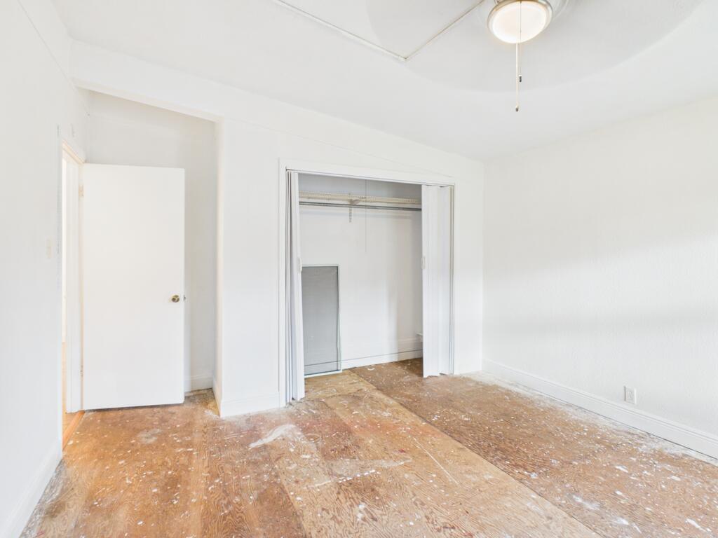 Empty bedroom with an open closet, white walls, and a worn bare wood floor.