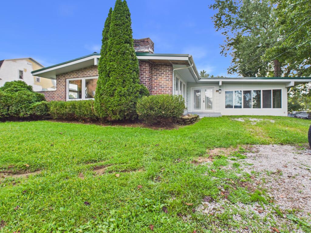 Single-story brick home with a white addition, landscaped shrubs, lawn, and gravel driveway.