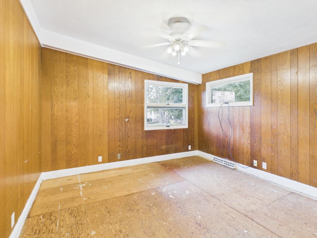 Vacant wood-paneled room with two windows, a ceiling fan, and worn plywood flooring.