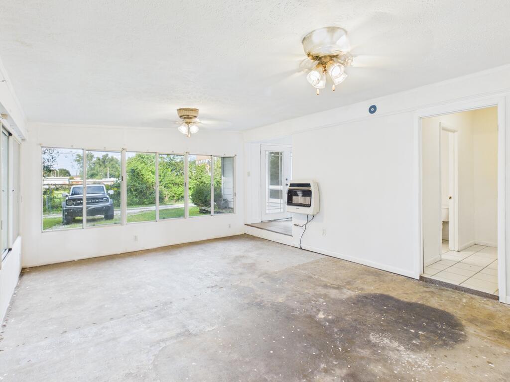 Empty living room with broad windows, a wall heater, and worn unfinished flooring.