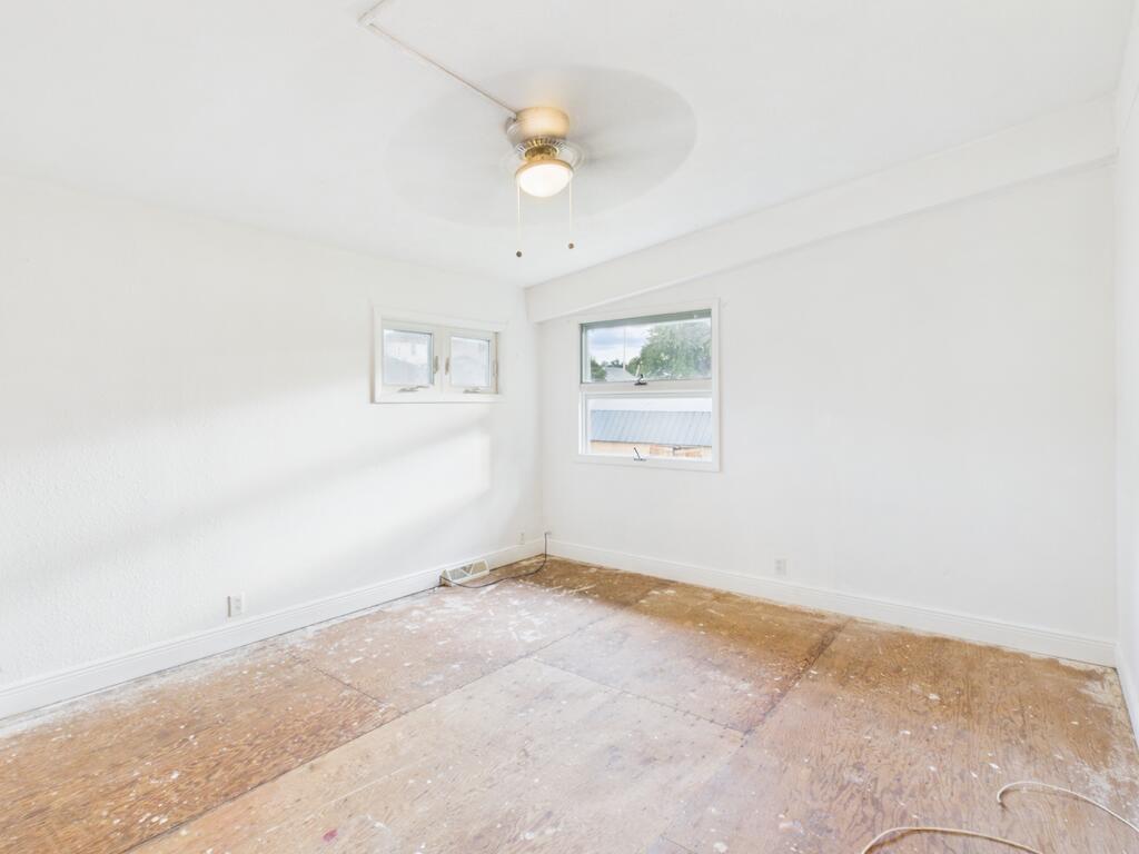 Empty room with white walls, two windows, a ceiling fan, and exposed plywood flooring.