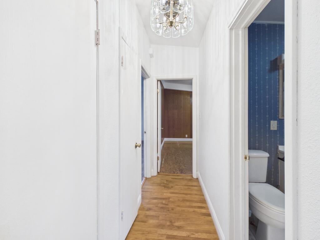 White hallway with wood flooring and chandelier, leading to a wood-paneled room beside a blue bathroom.