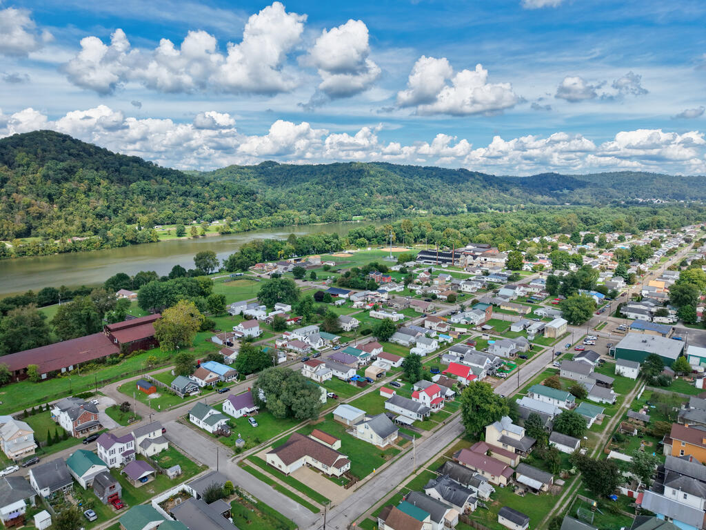 Aerial view of homes and streets beside a river, with wooded hills beyond.