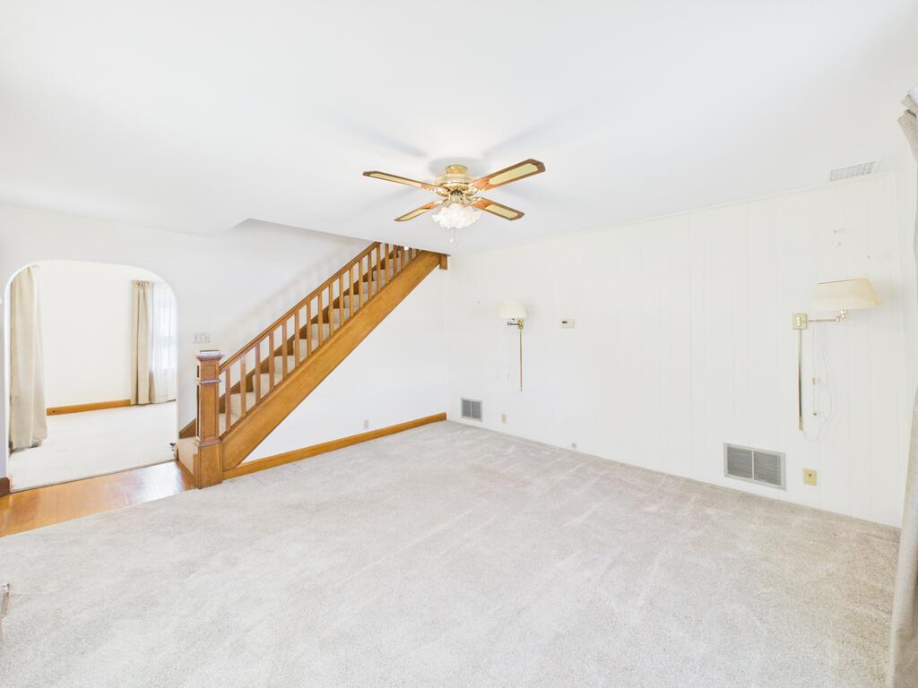 Empty living room with a wooden staircase, ceiling fan, and arched opening to an adjoining room.