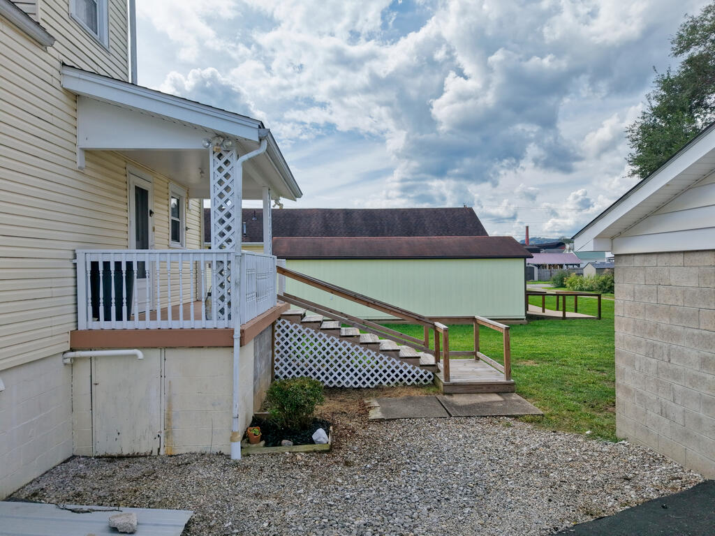 House porch with a wooden accessibility ramp, gravel yard, and nearby outbuildings.