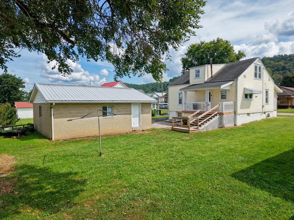 Light-colored two-story house with a raised front deck beside a detached block outbuilding and broad lawn.