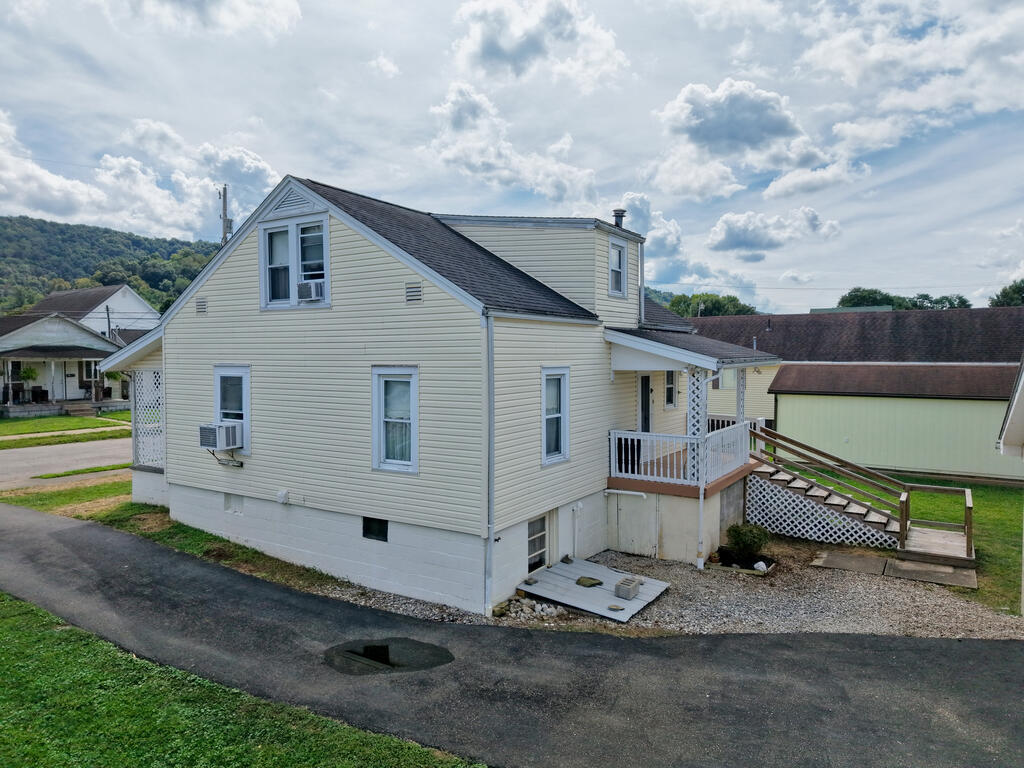Light-sided two-story house with a raised deck and exterior stairs beside a driveway.