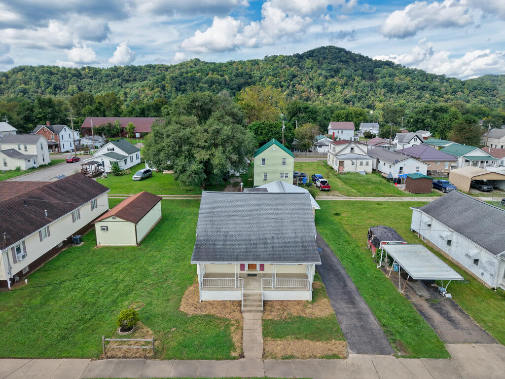 Aerial view of a house with a front porch, surrounded by lawns and neighboring homes beneath wooded hills.
