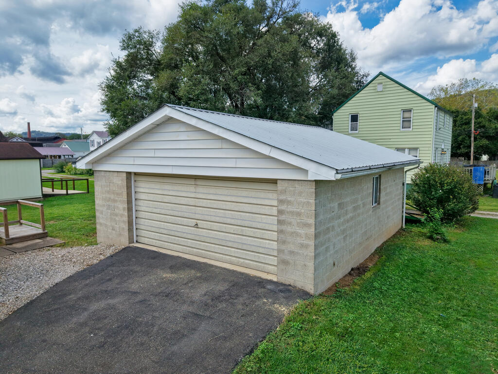 Detached block garage with a white metal roof and paved driveway, with a green house behind it.