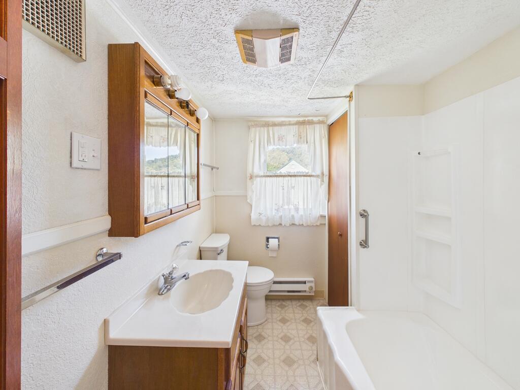 Bathroom with a long vanity, mirrored wooden cabinet, toilet, and bathtub beneath a curtained window.