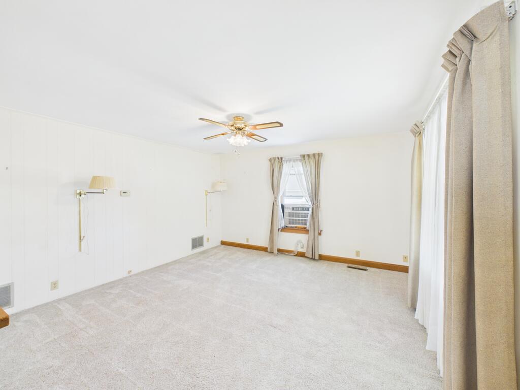 Empty living room with light carpet, beige curtains, and a ceiling fan.