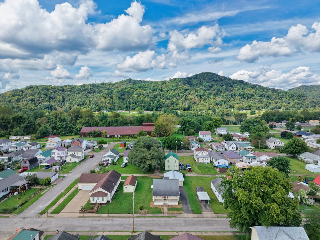 Aerial view of homes in a small town beneath wooded hills and a cloud-filled sky.