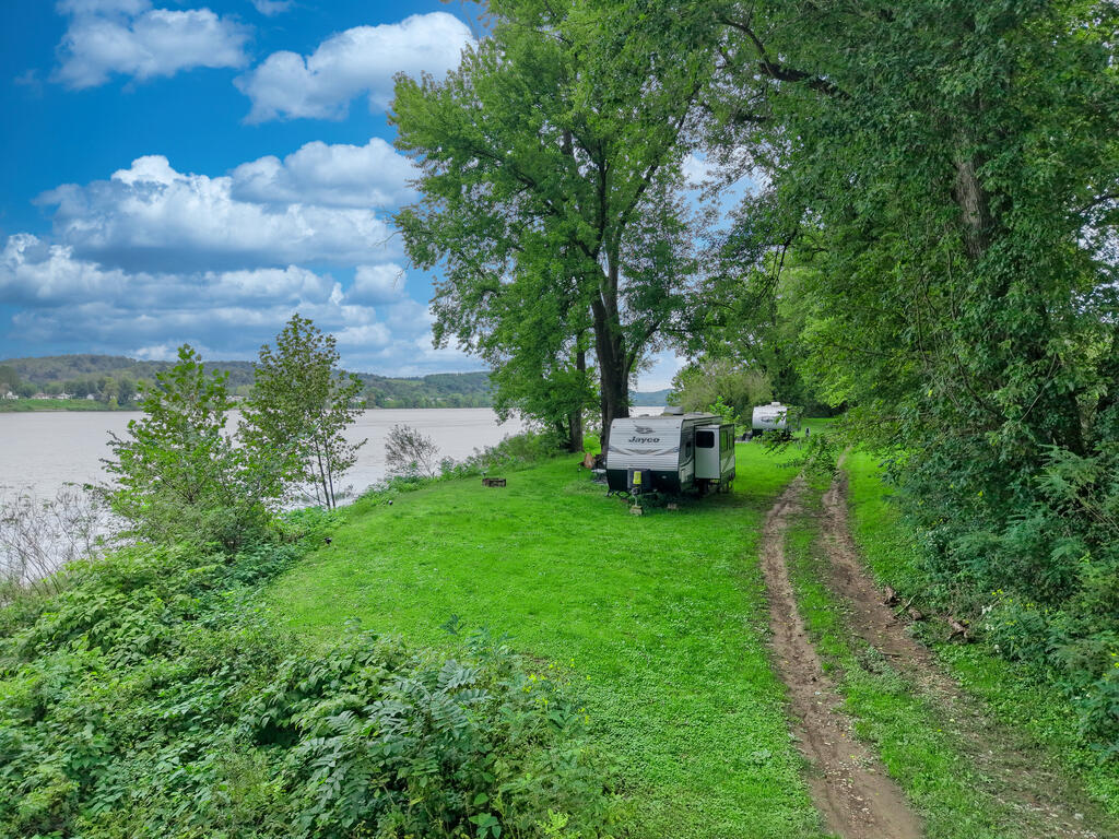 Grassy riverfront lot with a travel trailer, mature trees, and a dirt access track beside the water.