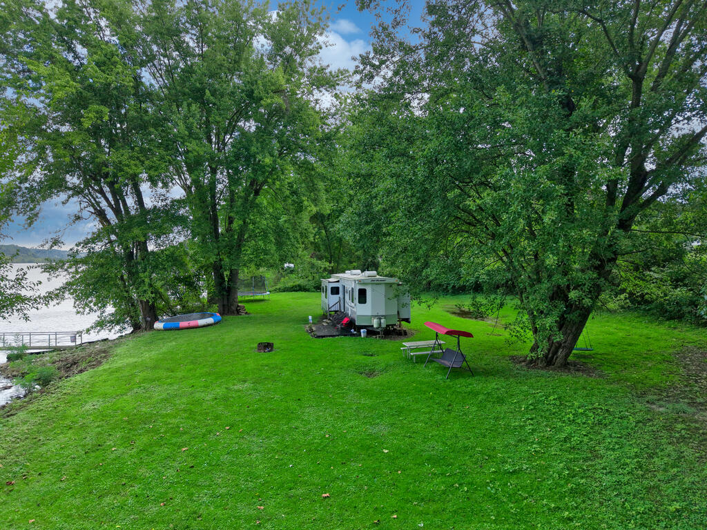 Riverfront lot with a shaded lawn, parked RV, outdoor chairs, and boat beside the water.