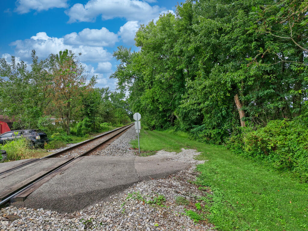 Grassy, wooded lot beside railroad tracks, with gravel and pavement in the foreground.