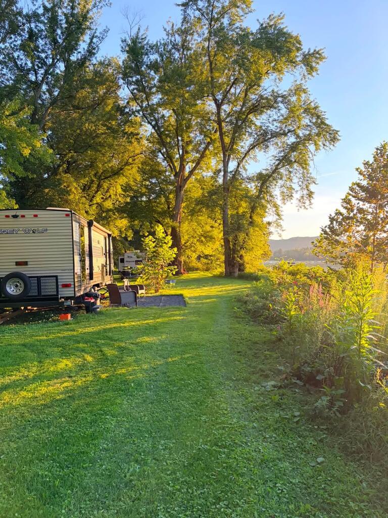 Grassy river lot with a travel trailer beneath tall autumn trees and distant hills.