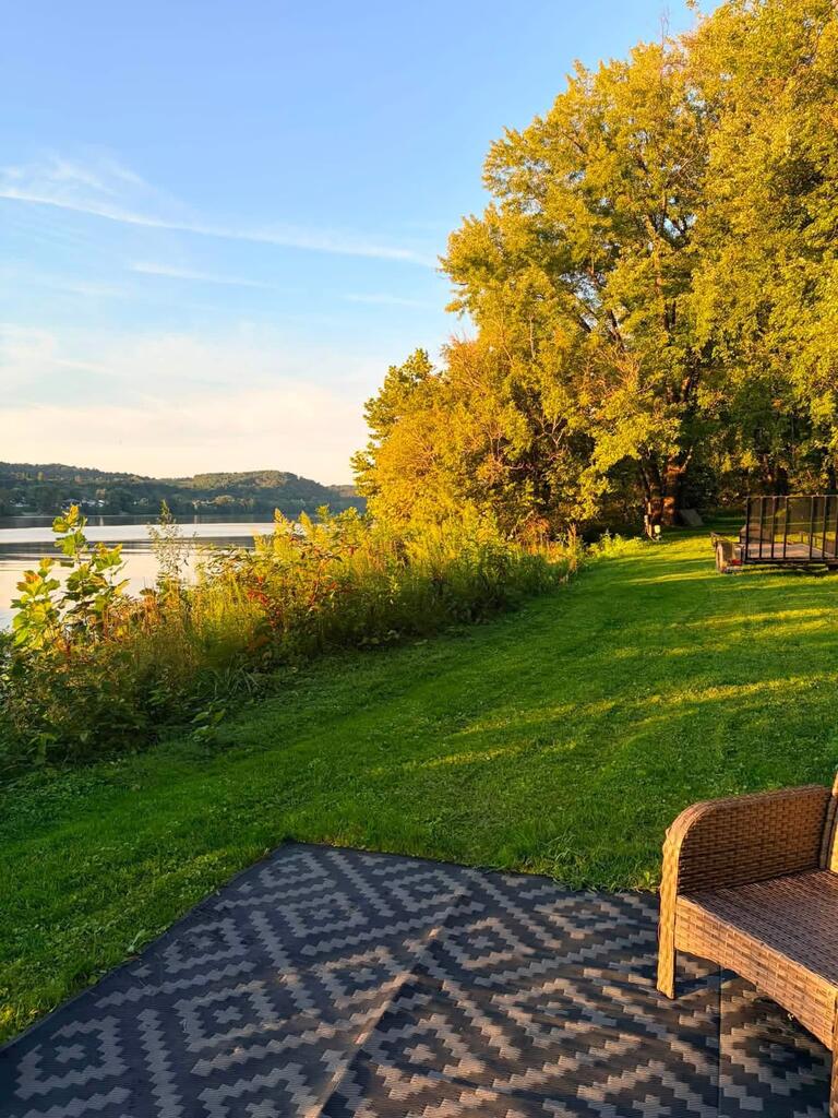 Riverfront lawn edged by golden autumn trees, with calm water beyond and a patio chair in the foreground.