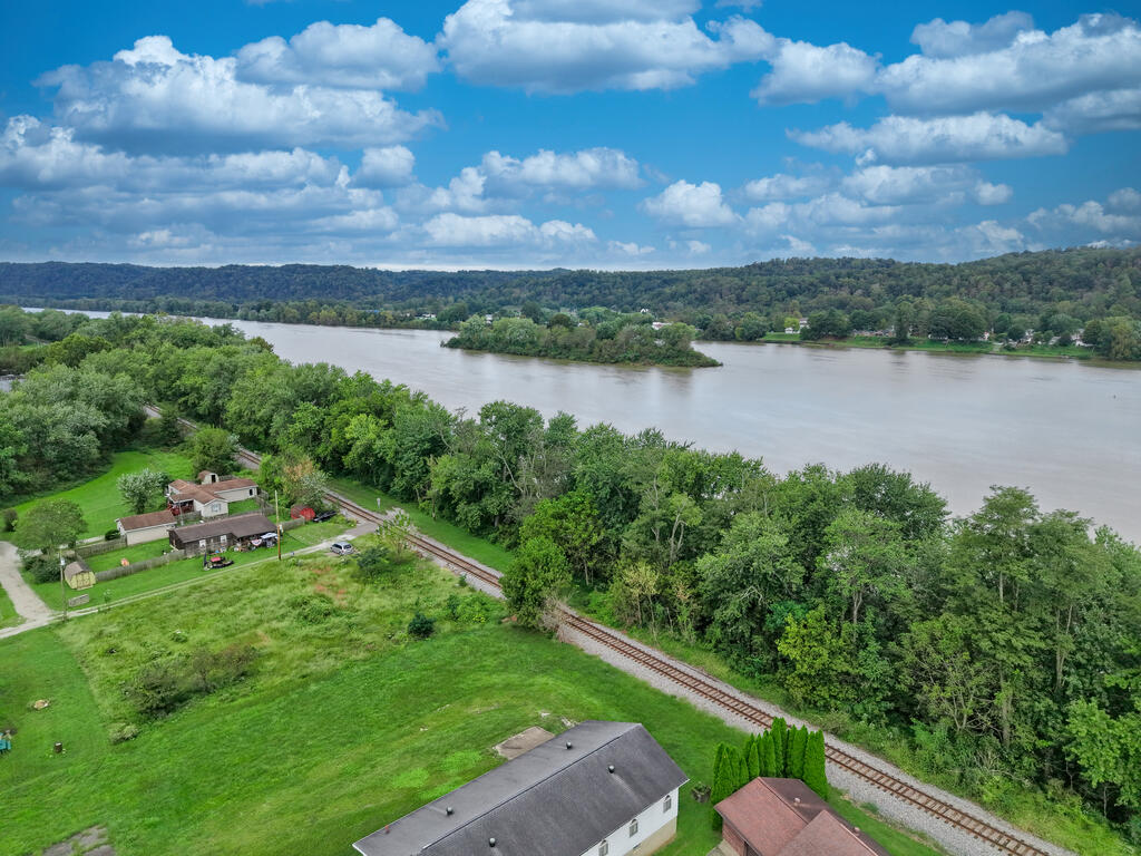 Aerial view of a green riverfront lot with railroad tracks and a broad river beyond.