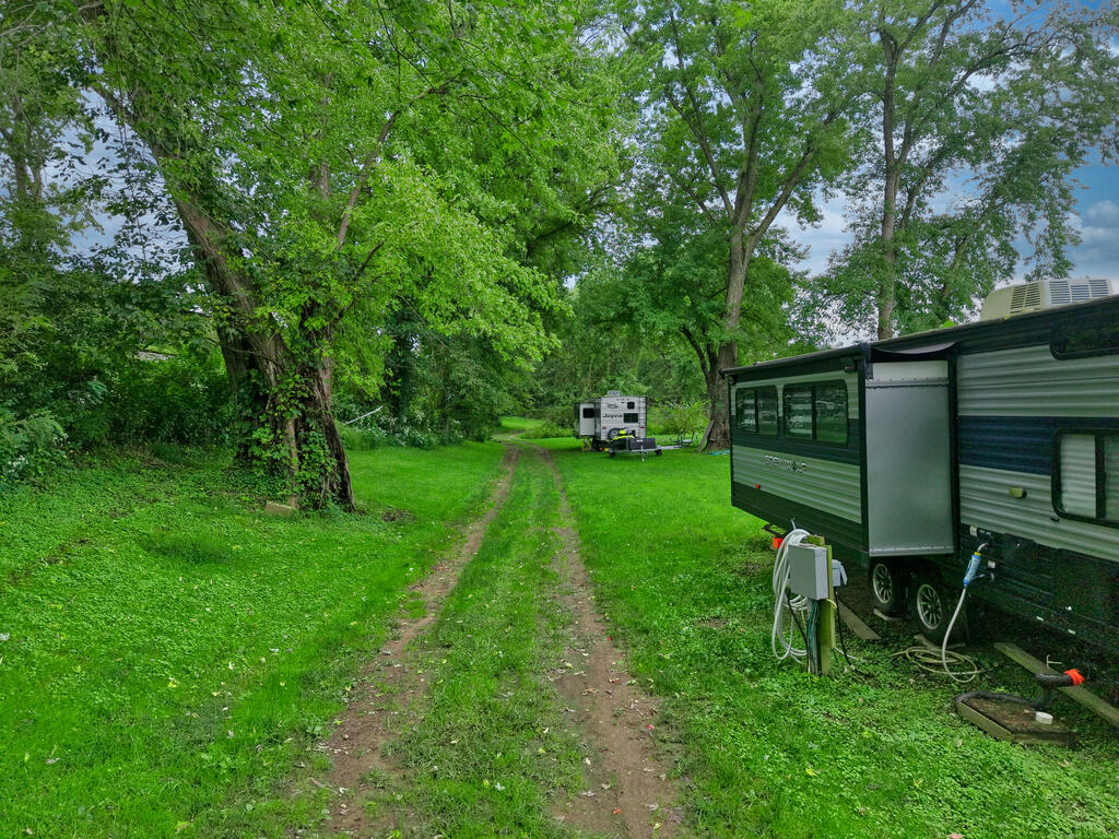 Grassy wooded lot with a dirt driveway, a camper ahead, and a travel trailer at right.