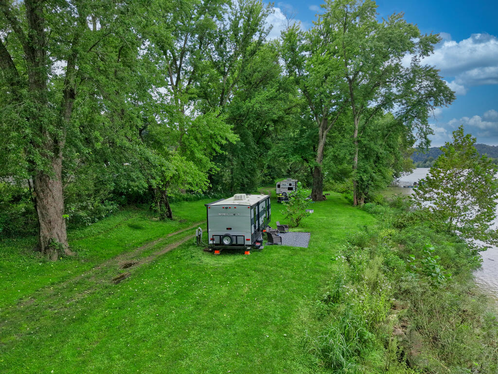 Grassy riverfront lot with mature trees, two parked campers, and a gravel area beside the river.