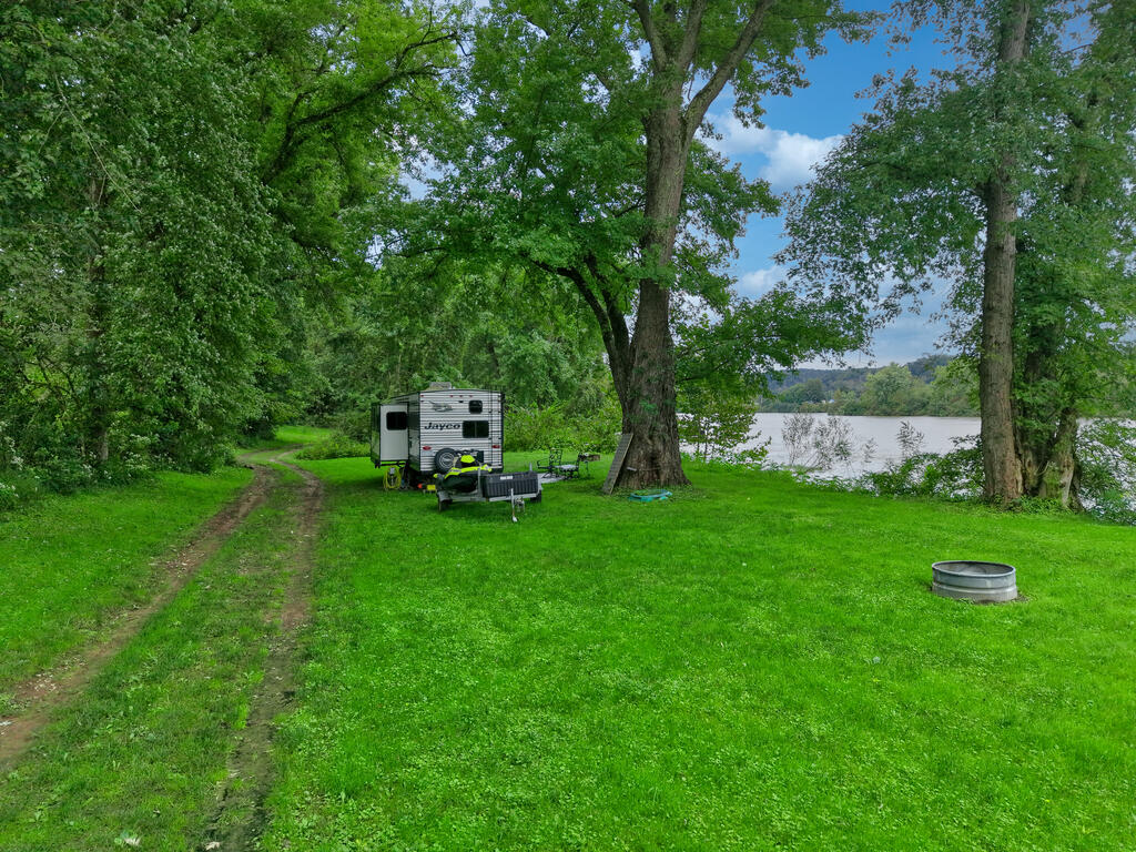 Grassy riverside lot shaded by mature trees, with a travel trailer and metal fire ring.