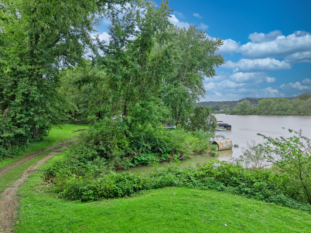 Grassy riverfront lot with a dirt access path, mature trees, and boats along the water.