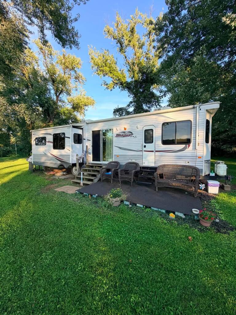 White travel trailer on a grassy, tree-shaded lot, with outdoor seating in front.