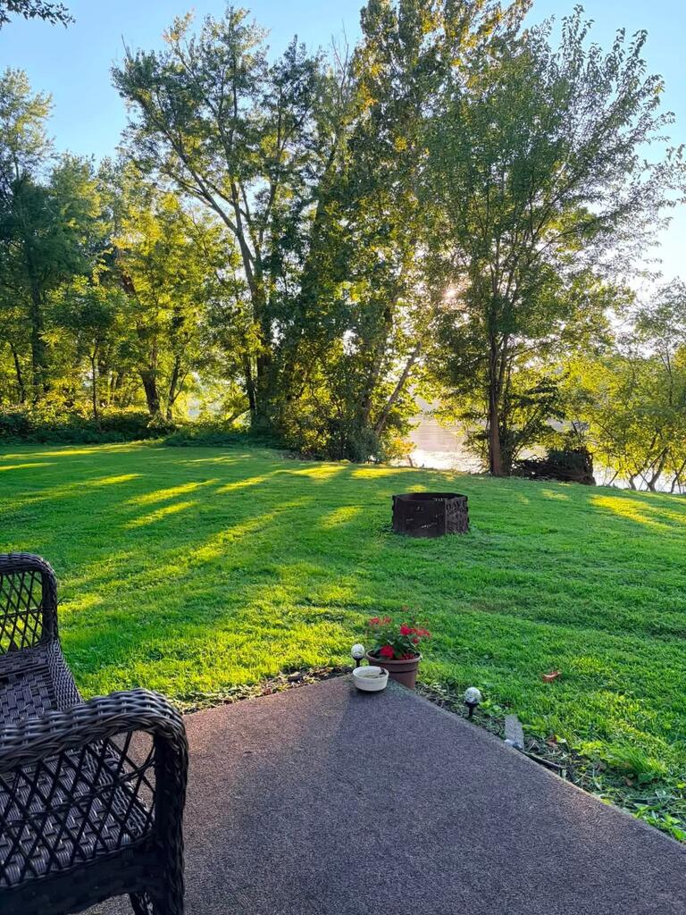 Riverfront lawn shaded by mature trees, with a paved patio, wicker chair, and fire pit.