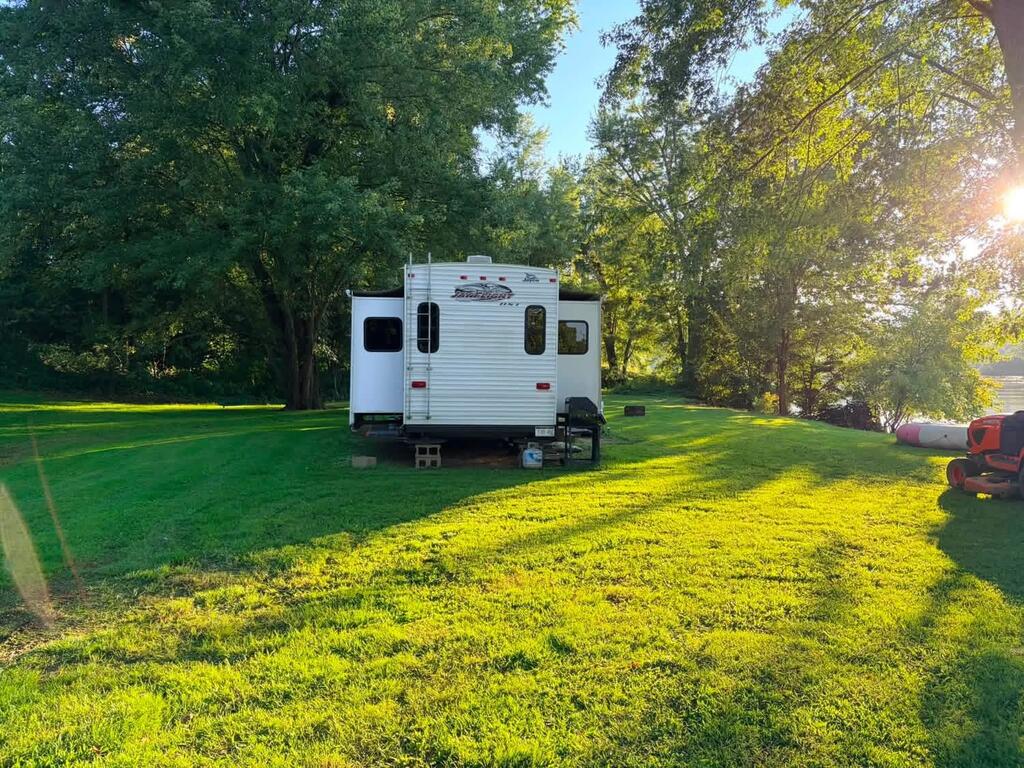Grassy river lot with a travel trailer beneath mature trees and water visible at the edge.