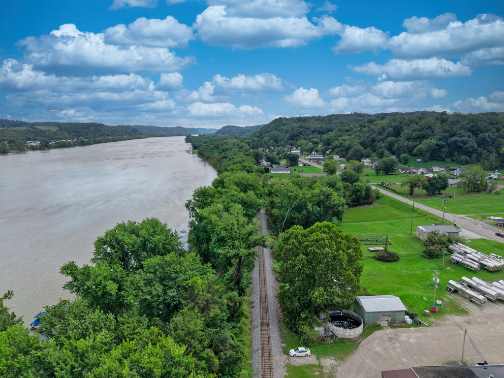 Aerial view of a riverfront lot beside railroad tracks, with wooded hills and homes beyond.