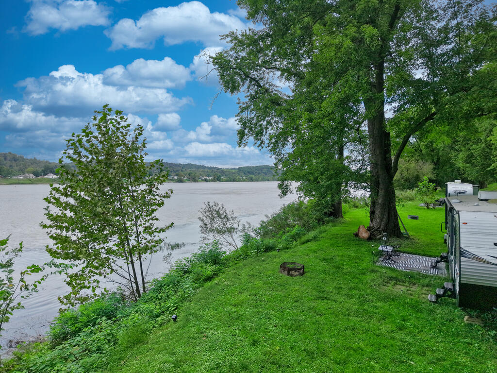 Riverfront lot with a grassy bank, mature trees, fire ring, and camper beside the river.