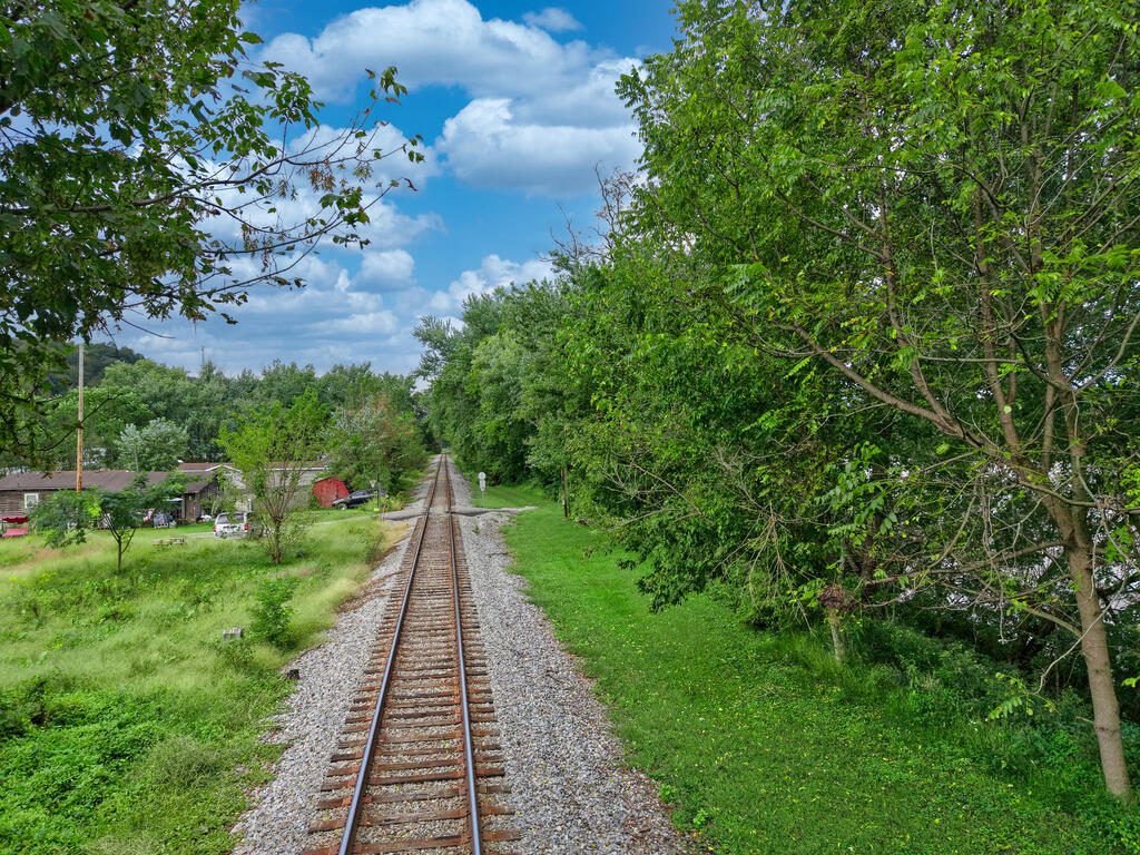 Tree-lined grassy lot beside railroad tracks, with small buildings in the distance.