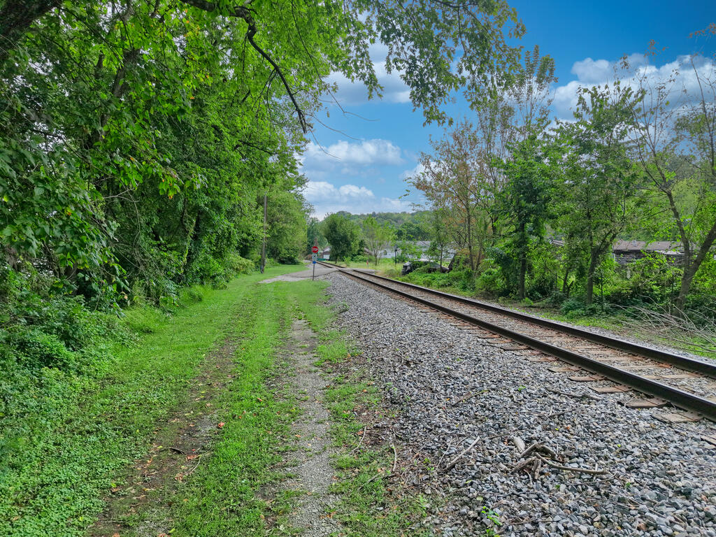 Wooded grassy lot beside railroad tracks, with a gravel path and leafy trees.