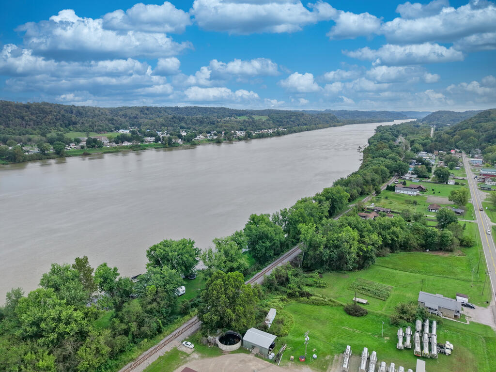 Aerial view of a riverfront lot with a tree-lined shore, railroad tracks, and nearby homes.