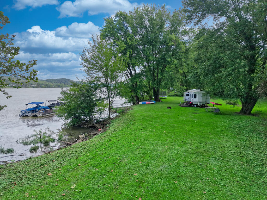 Grassy riverfront lot with mature trees, a moored boat, and a camper trailer.