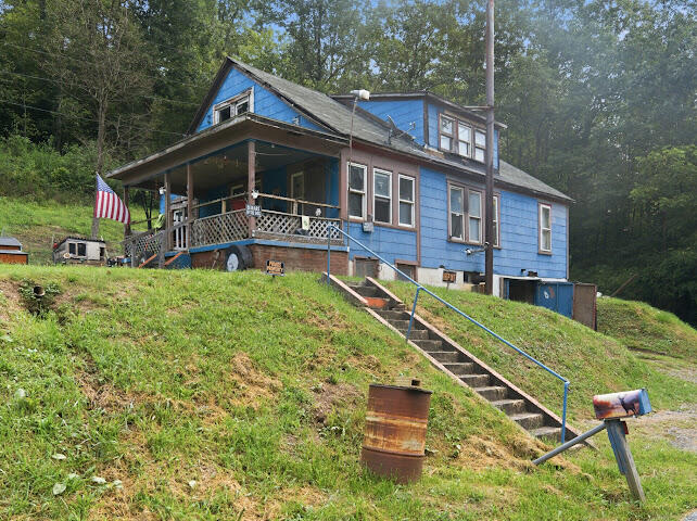 Blue two-story house on a grassy hillside, with a covered porch and stairs leading down to the road.