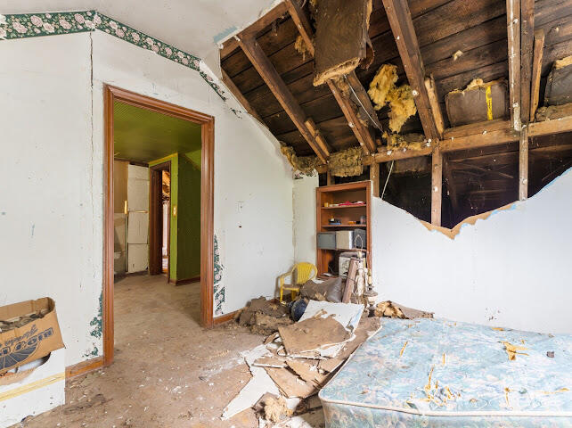 Damaged bedroom with exposed rafters, scattered debris, and a mattress.
