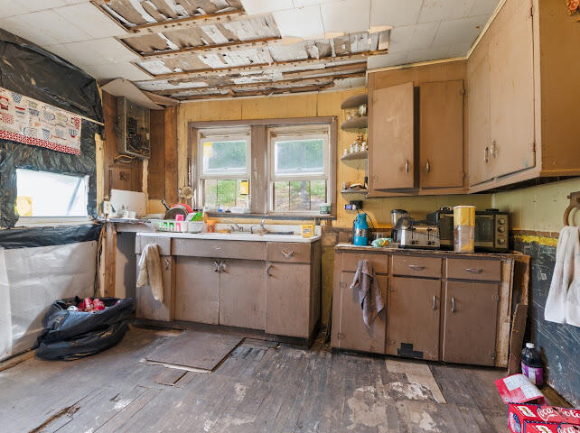Kitchen with worn cabinets, a sink beneath two windows, and damaged ceiling and flooring.