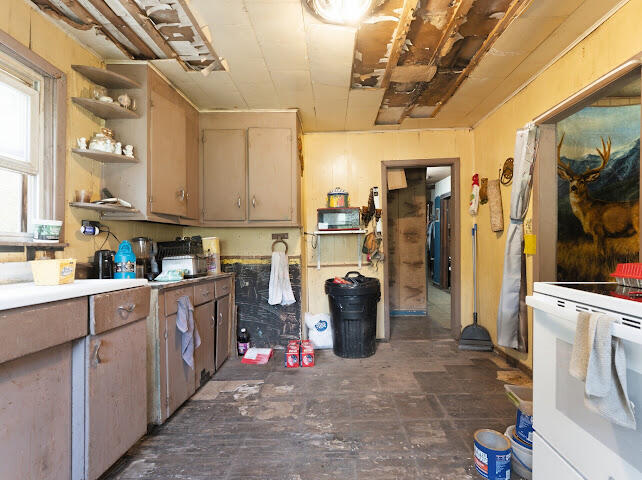 Worn kitchen with left-side cabinets and sink, appliances, and a damaged exposed ceiling.