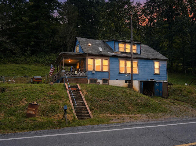 Blue-sided house with warmly lit windows, a covered porch, and hillside steps.