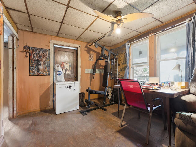 Multipurpose room with exercise equipment, a desk by two windows, and a chest freezer.